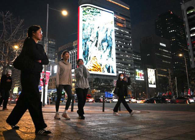 People walk in front of digital billboards near Gwanghwamun Square in Seoul on April 1, 2026. Hopes of making downtown Seoul dazzle more than Times Square have hit a setback with new guidelines to dim the massive digital billboards that light up the South Korean capital after a barrage of complaints. (Photo by Jung Yeon-je / AFP)