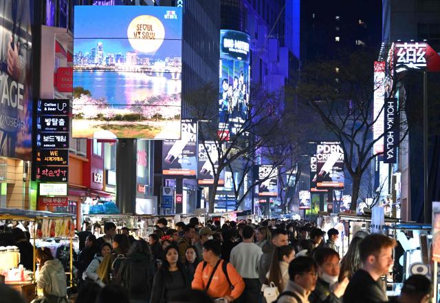 People walk beneath digital billboards at Myeongdong shopping district in Seoul on April 1, 2026. Hopes of making downtown Seoul dazzle more than Times Square have hit a setback with new guidelines to dim the massive digital billboards that light up the South Korean capital after a barrage of complaints. (Photo by Jung Yeon-je / AFP)