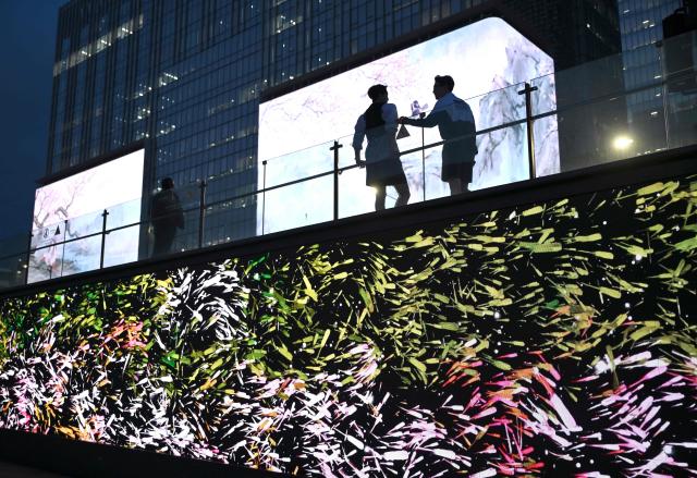 TOPSHOT - People stand in front of digital billboards at Gwanghwamun Square in Seoul on April 1, 2026. Hopes of making downtown Seoul dazzle more than Times Square have hit a setback with new guidelines to dim the massive digital billboards that light up the South Korean capital after a barrage of complaints. (Photo by Jung Yeon-je / AFP)
