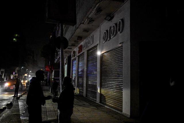 People stand near shops closed early under a government-ordered curfew aimed at reducing energy costs in downtown Cairo on April 2, 2026. Egypt has ordered shops, restaurants and shopping malls to close from 9:00 pm from April 3, hoping to curb energy bills that have more than doubled because of the Iran war. Prime Minister Mostafa Madbouly announced the curfew and said it would last for a month initially. (Photo by Khaled DESOUKI / AFP)