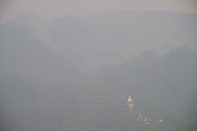 A general view shows a white Buddha statue shrouded in heavy pollution as smoke (top L) rises from a hillside wildfire in Pai on April 1, 2026, west of Thailand's second-largest city Chiang Mai. Seasonal agricultural burning, forest fires and weather patterns produce an annual pollution season across much of Southeast Asia. But parts of northern Thailand are seeing haze that even hardened locals say is exceptional. (Photo by ANTHONY WALLACE / AFP) / To go with 'Thailand-Pollution-Environment-Fire-Health' Reportage by Chayanit ITTHIPONGMAETEE