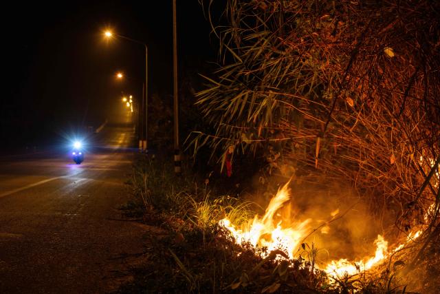 A wildfire is seen along a road as a motorist drives past in Chiang Mai on April 1, 2026. Seasonal agricultural burning, forest fires and weather patterns produce an annual pollution season across much of Southeast Asia. But parts of northern Thailand are seeing haze that even hardened locals say is exceptional. (Photo by ANTHONY WALLACE / AFP) / To go with 'Thailand-Pollution-Environment-Fire-Health' Reportage by Chayanit ITTHIPONGMAETEE