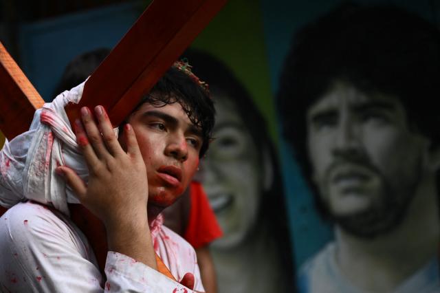 A youngster enacts the Passion of Jesus Christ with a mural depicting Argentina's football legend Diego Maradona in the background, during a procession marking the Way of the Cross on Good Friday at the Padre Carlos Mujica shantytown (known as Villa 31) in Buenos Aires on April 3, 2026. (Photo by Luis ROBAYO / AFP)