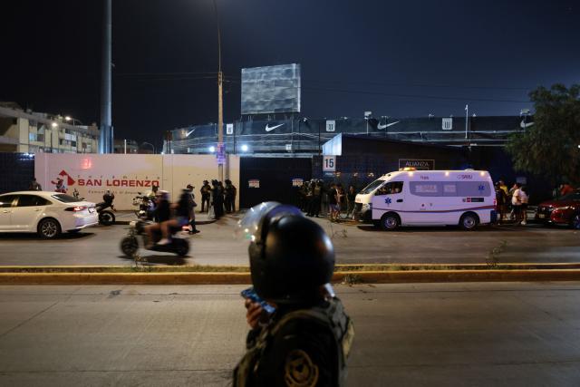 TOPSHOT - Police officers stand guard at the entrance of the Alejandro Villanueva Stadium while relatives wait for news after an accident in the stands where Alianza Lima's fans were cheering for their team on the eve of the match against Universitario in Lima on April 3, 2026. At least one person was killed and 47 others were injured, including three minors, in “an incident” that occurred Friday at Alianza Lima's stadium in Peru during a flag-waving rally in support of the team ahead of the Peruvian football derby, the Ministry of Health reported. (Photo by Connie FRANCE / AFP)
