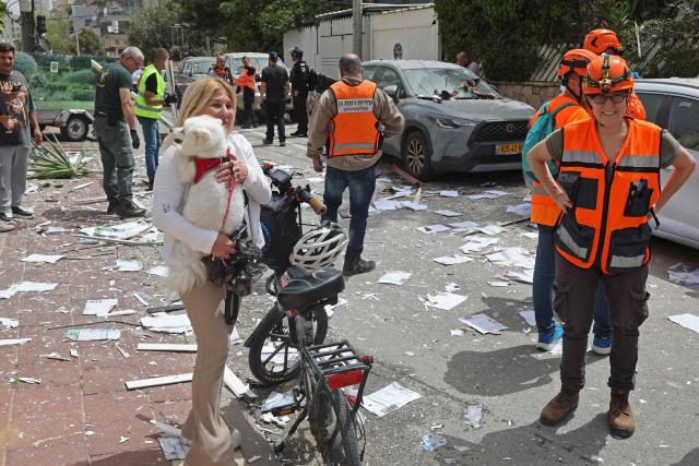 A woman carries her dog next to Israel's first responders at the site of an Iranian strike that hit a residential neighbourhood in Ramat Gan on April 4, 2026. The Israeli army said on April 4 its air defences were working to down missiles fired from Iran, which medics reported wounded one person. (Photo by Jack GUEZ / AFP) / 
