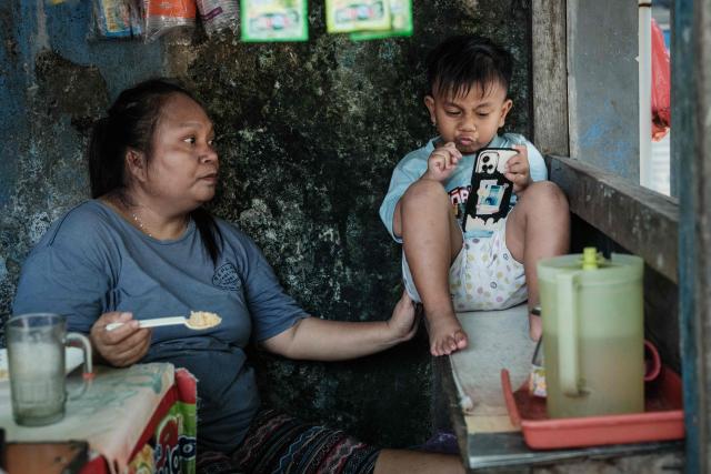 A three-and-a-half-year-old boy uses a smartphone as his mother waits to feed him at a food stall in Jakarta on April 4, 2026. (Photo by YASUYOSHI CHIBA / AFP)