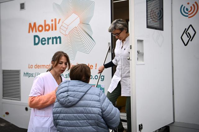 Dermatologist Blandine De Carrere (R) steps out of an itinerant truck as medical assistant and truck driver Lea Fougerouge (L) speaks with a patient ahead of an appointment as part of the Mobil Derm project supported by the Societe Francaise de Dermatologie (French Dermatology Society) at a parking lot in Lafox, south-western France, on April 2, 2026. As in the rest of France, patients suffering from skin diseases are facing significant delays in receiving care in Nouvelle-Aquitaine, with loss of chances in the most serious cases. In Lafox, 25 patients a day enter the few square meters of the mobile clinic, where a dermatologist from the Basque Country has come for two days to carry out consultations in her free time  a pioneering system in France intended to compensate for the shortage of dermatologists and address medical deserts. (Photo by Christophe ARCHAMBAULT / AFP)