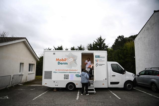 Medical assistant and truck driver Lea Fougerouge speaks with a patient ahead of an appointment with a dermatologist in an itinerant truck as part of the Mobil Derm project supported by the Societe Francaise de Dermatologie (French Dermatology Society) at a parking lot in Lafox, south-western France, on April 2, 2026. As in the rest of France, patients suffering from skin diseases are facing significant delays in receiving care in Nouvelle-Aquitaine, with loss of chances in the most serious cases. In Lafox, 25 patients a day enter the few square meters of the mobile clinic, where a dermatologist from the Basque Country has come for two days to carry out consultations in her free time  a pioneering system in France intended to compensate for the shortage of dermatologists and address medical deserts. (Photo by Christophe ARCHAMBAULT / AFP)