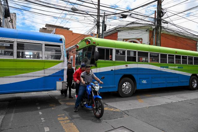 A motorcyclist rides between parked buses as part of a protest by transport operators in front of the Metropolitan Cathedral in Guatemala City on April 6, 2026. Transport operators call for the Guatemalan government's help in the face of rising fuel and oil derivative prices due to the Middle East crisis. (Photo by Johan ORDÓÑEZ / AFP)