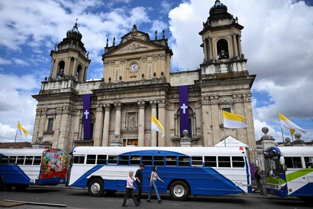 People walk past parked buses as part of a protest by transport operators in front of the Metropolitan Cathedral in Guatemala City on April 6, 2026. Transport operators call for the Guatemalan government's help in the face of rising fuel and oil derivative prices due to the Middle East crisis. (Photo by Johan ORDÓÑEZ / AFP)
