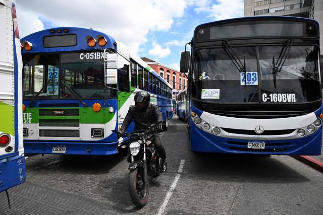 A motorcyclist rides between parked buses as part of a protest by transport operators in front of the Metropolitan Cathedral in Guatemala City on April 6, 2026. Transport operators call for the Guatemalan government's help in the face of rising fuel and oil derivative prices due to the Middle East crisis. (Photo by Johan ORDÓÑEZ / AFP)