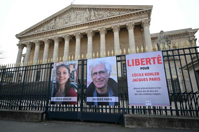 (FILES) This photograph shows the newly displayed portraits of French national Cecile Kohler (L) currently imprisoned in Iran with her partner French national Jacques Paris (C) along with a placard reading "Freedom for Cecile Kohler and Jacques Paris arbitrarily detained in Iran for over two years in appalling conditions" outside the Palais Bourbon, France's National Assembly, following their meeting with the President of France's National Assembly, in Paris, on March 25, 2025. Former detainees Cecile Kohler and Jacques Paris "are free and on their way to France", France's President Emmanuel Macron announced on April 7, 2026. (Photo by Bertrand GUAY / AFP)