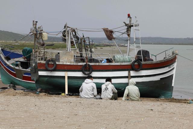 Men sit on the sand next to a fishing boat in Chelihat, Kenitra region on April 7, 2026. (Photo by Abdel Majid BZIOUAT / AFP)