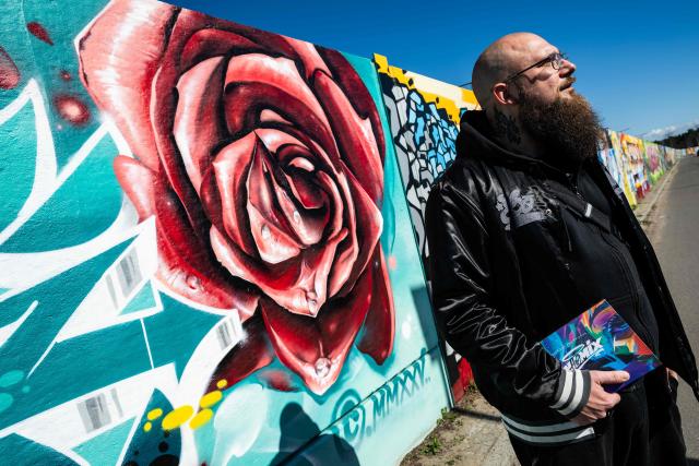 German rapper and street artist Akte One (Mark Marquardt) poses next to the recently unveiled "Sued Ost Galerie" open air collaborative graffiti art exhibition in Berlin on April 8, 2026. The 450-metre-long noise protection wall located in the capital's south east features mural art by some 20 artists collaborating around Akte one who initiated the project in 2025. (Photo by John MACDOUGALL / AFP)