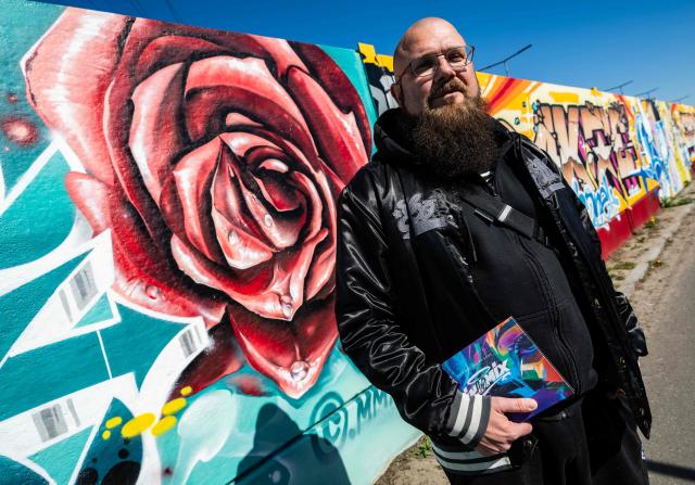German rapper and street artist Akte One (Mark Marquardt) poses next to the recently unveiled "Sued Ost Galerie" open air collaborative graffiti art exhibition in Berlin on April 8, 2026. The 450-metre-long noise protection wall located in the capital's south east features mural art by some 20 artists collaborating around Akte one who initiated the project in 2025. (Photo by John MACDOUGALL / AFP)