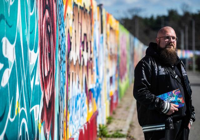 German rapper and street artist Akte One (Mark Marquardt) poses next to the recently unveiled "Sued Ost Galerie" open air collaborative graffiti art exhibition in Berlin on April 8, 2026. The 450-metre-long noise protection wall located in the capital's south east features mural art by some 20 artists collaborating around Akte one who initiated the project in 2025. (Photo by John MACDOUGALL / AFP)