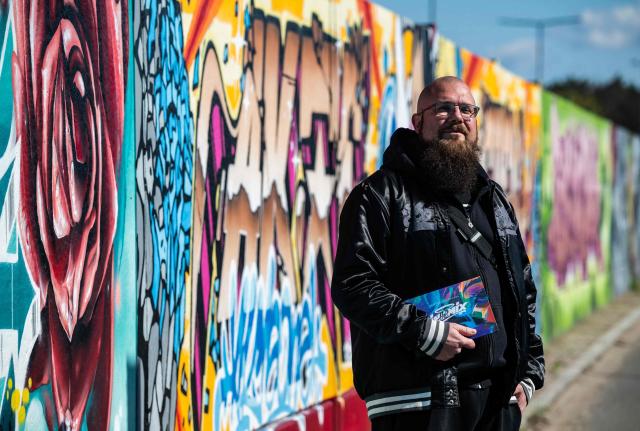 German rapper and street artist Akte One (Mark Marquardt) poses next to the recently unveiled "Sued Ost Galerie" open air collaborative graffiti art exhibition in Berlin on April 8, 2026. The 450-metre-long noise protection wall located in the capital's south east features mural art by some 20 artists collaborating around Akte one who initiated the project in 2025. (Photo by John MACDOUGALL / AFP)
