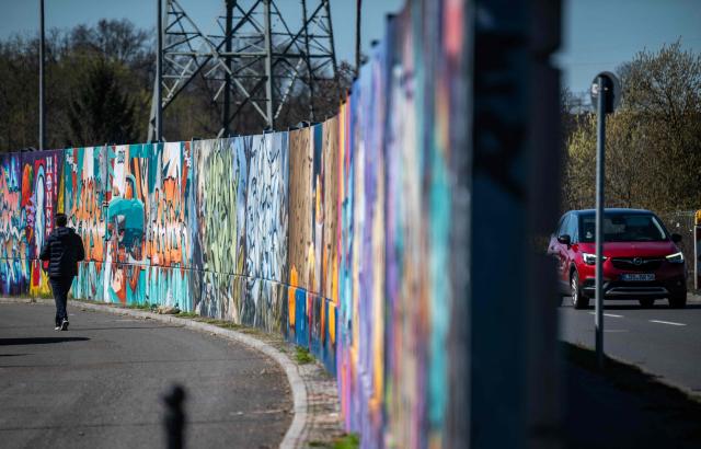 A man walks along the recently unveiled "Sued Ost Galerie" open air collaborative graffiti art exhibition in Berlin on April 8, 2026. The 450-metre-long noise protection wall located in the capital's south east features mural art by some 20 artists collaborating around German rapper and street artist Akte One (Mark Marquardt) who initiated the project in 2025. (Photo by John MACDOUGALL / AFP)