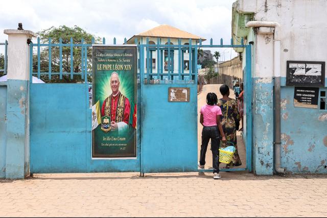 People pass through the gates of La Retraite College, which displays a portrait of Pope Leo XIV, in Yaounde on April 8, 2026. (Photo by Daniel BELOUMOU OLOMO / AFP)