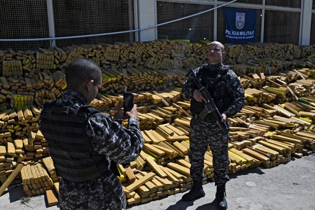 A police officer poses for a photo next to 48 tons of seized marijuana presented at the City of Police in Rio de Janeiro, Brazil on April 8, 2026, after they were uncovered by chance by a sniffer dog during a regular operation against criminal factions. During the operation in the Complexo da Mare, a large complex of favelas in the north of the city, a dog named Hulk indicated there was something suspicious under an abandoned water tank. An officer "discovered a bunker inside where this entire quantity of drugs had been hidden," said Lieutenant Colonel Luciano Pedro, commander of the Canine Operations Battalion. (Photo by Pablo PORCIUNCULA / AFP)