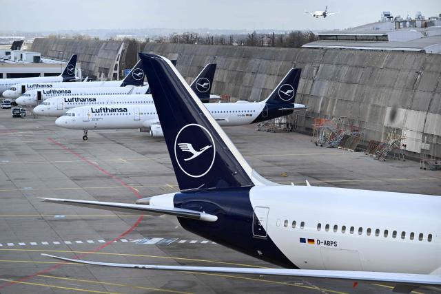 (FILES) The logo of German airline Lufthansa is seen on vertical stabilisers of aircraft parked on the tarmac while in the background an airplace is seen in flight at the international Frankfurt Airport, Frankfurt am Main, western Germany, on March 12, 2026. Cabin crew at German airline Lufthansa will go on strike on April 10, 2026 over an ongoing labour dispute, their union said. (Photo by Kirill KUDRYAVTSEV / AFP)