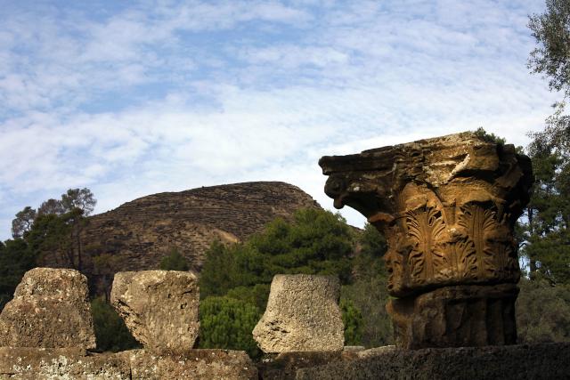 (FILES) A part of an ancient column in the archeological site of Olympia stands in front of the charred ancient hill of Kronos, 24 November 2007. Greek and international scientists visited the World Heritage site to deliver funds for reconstruction of the area. A large replanting effort is under way ahead of March 25, when the flame-lighting ceremony for the Beijing Olympics will be held at the birthplace of the ancient games. From 2022 to 2025, scientists at the National University of Athens and the National Hellenic Research Foundation examined past and present climate and geological conditions at the 19 sites, looking at prior damage to help determine future vulnerability to extreme events. The sites under scrutiny include Olympia, habitually threatened by forest fires, the ancient theatre of Delphi, where rockslides are a concern. (Photo by Aris MESSINIS / AFP)