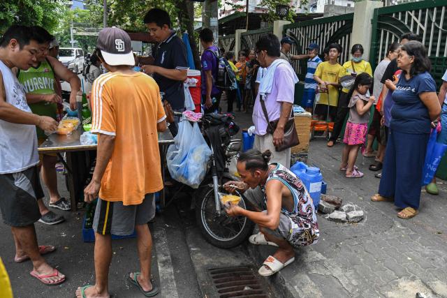 People eat their meal sold by a vendor (L) while residents (R) queue up outside a government-sponsored rolling store selling cheap rice near a public market in Manila on April 10, 2026. (Photo by Ted ALJIBE / AFP)