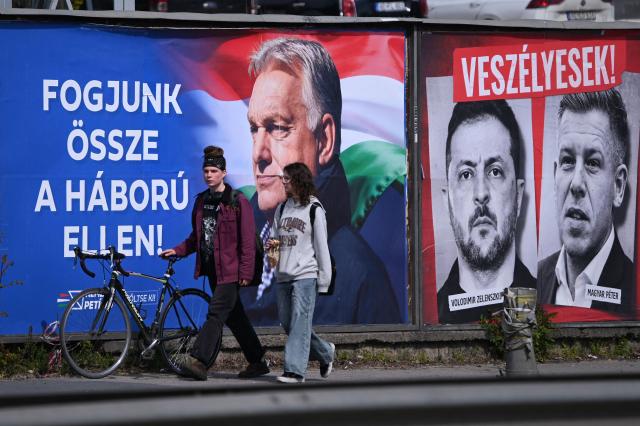 A young couple walk in front of billboards, featuring Hungarian Prime Minister Viktor Orban (L) with a text, reading 'Come together against the war' as portrait of Ukraine's President Volodymyr Zelensky and Hungarian opposition leader Peter Magyar with the text reading, 'They are dangerous',(top) and 'Let's stop them, just Fidesz (governor party)(R)' are seen in Budapest's 11th district on April 10, 2026, two days before the elections. The "poster war" has entered an ever more intense phase in the run-up to the election campaign. Cities and roads are flooded with political advertisements. The elections will be held in Hungary on April 12. (Photo by ATTILA KISBENEDEK / AFP)