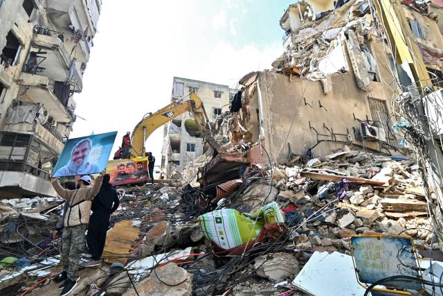 This photograph taken on April 10, 2026, during a media tour organized by Hezbollah’s media office shows a man holding a poster of slain Lebanese Hezbollah leader Hassan Nasrallaha as a digger clears the rubble of a building in Beirut's Hay al-Selloum neighbourhood, that was targetted in an Israeli strike earlier this week. The Israeli military said on April 10 that it had "dismantled" more than 4,300 Hezbollah sites in Lebanon since fighting with the militant group began last month. (Photo by FADEL itani / AFP) / 
