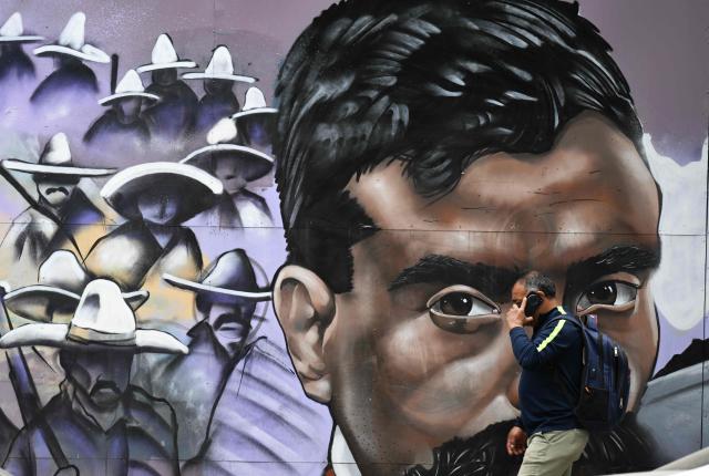 A man uses his mobile phone in front of a mural by Kolore Fuentes depicting the iconic hero of the Mexican Revolution, Emiliano Zapata in Mexico City on April 10, 2026. Mexico commemorates the 107th anniversary of Zapata's death. (Photo by Yuri CORTEZ / AFP)