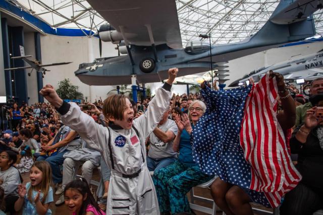 A young boy wearing an astronaut costume cheers next to a woman waving a flag as they watch a live broadcast of the return of the Artemis II crew members to Earth at the San Diego Air and Space Museum during a watch party for the crew's splash down in the Pacific Ocean, in San Diego, California, on April 10, 2026. The NASA spacecraft carrying four astronauts splashed down as planned Friday off the California coast, capping the US space agency's successful crewed test mission around the Moon, the first such flyby in more than 50 years. Mission commander Reid Wiseman reported that the crewmembers -- himself along with Christina Koch, Victor Glover and Jeremy Hansen -- were "stable" and "green." (Photo by Apu GOMES / AFP)