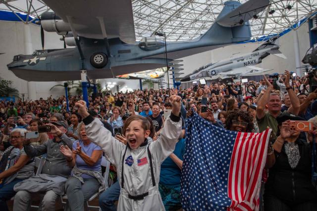 A young boy wears an astronaut costume next to a woman waving a flag as they watch a live broadcast of the return of the Artemis II crew members to Earth at the San Diego Air and Space Museum during a watch party for the crew's splash down in the Pacific Ocean, in San Diego, California, on April 10, 2026. The NASA spacecraft carrying four astronauts splashed down as planned Friday off the California coast, capping the US space agency's successful crewed test mission around the Moon, the first such flyby in more than 50 years. Mission commander Reid Wiseman reported that the crewmembers -- himself along with Christina Koch, Victor Glover and Jeremy Hansen -- were "stable" and "green." (Photo by Apu GOMES / AFP)