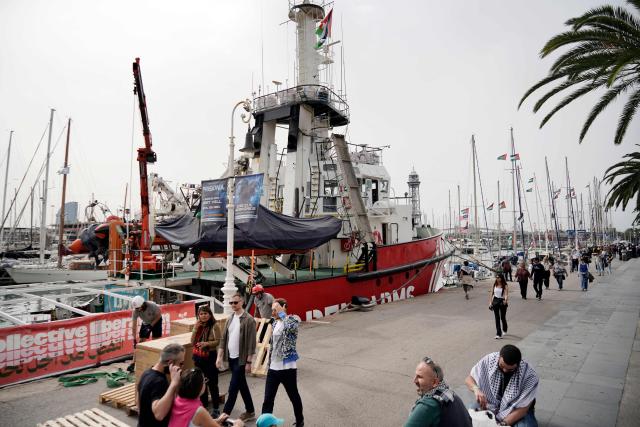 People walk past the NGO ship Open Arms, moored next to other vessels of a new humanitarian flotilla bound for the Gaza Strip at the port of Barcelona on April 11, 2026, on the eve of their departure. A new flotilla of more than 80 boats and 1,000 international pro-Palestinian activists, who attempted to reach Gaza last year, is preparing for a new mission to the devastated territory from Barcelona tomorrow. Israel's response to the Global Sumud Flotilla's first journey —intercepting the boats and arresting the activists— sparked international condemnation. (Photo by Manaure Quintero / AFP)