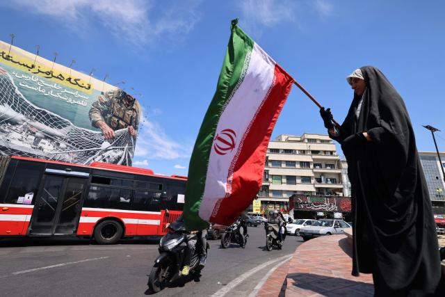 A woman waves Iran's national flag in front of a giant billboard reading 'The Strait of Hormuz remains closed' at the Revolution Square in Tehran on April 12, 2026. Iran's parliament speaker Mohammad Bagher Ghalibaf, who was part of peace talks with the United States this weekend, said on April 12 that Washington was "unable" to win Tehran's trust during the discussions. (Photo by ATTA KENARE / AFP) / 