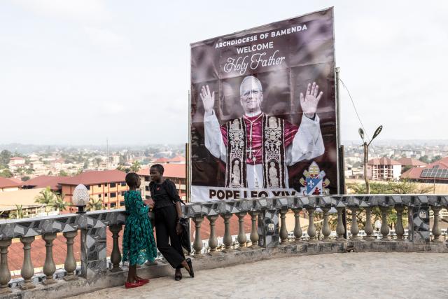 Young girls stand next to a billboard welcoming Pope Leo XIV ahead of his visit during Sunday Mass at Saint Joseph Metropolitan Cathedral in Bamenda, on April 12, 2026. (Photo by PATRICK MEINHARDT / AFP)