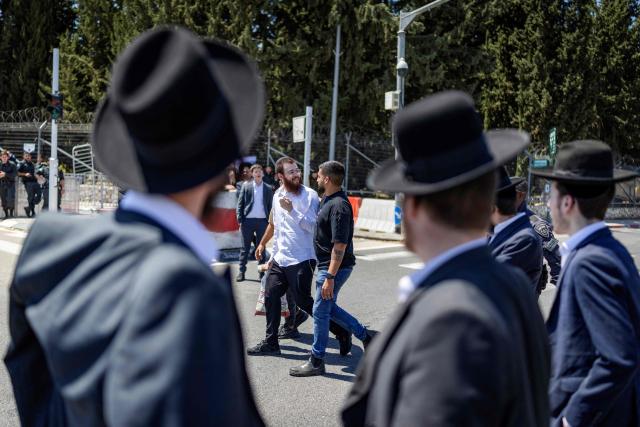 Israeli police escort an ultra-Orthodox Jewish man (C) into recruitment center as others, who are largely exempt from military service, gather during a protest against conscription at the entrance to Tel Hashomer, the central recruitment centre of the Israeli Army, in Ramat-Gan on April 12, 2026. Israel’s Prime Minister, who depends on his ultra-Orthodox party allies to maintain power, has employed a range of tactics to delay the adoption of legislation that would allow the enlistment of ultra-Orthodox Jews, who are largely exempt from military service. Under a ruling established at the country’s creation in 1948—when the ultra-Orthodox community was small—men who devote themselves full-time to the study of sacred Jewish texts receive a de facto exemption. (Photo by Ilia YEFIMOVICH / AFP) / 