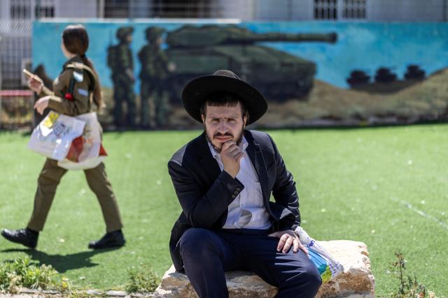 A female Israeli conscript walks past as ultra-Orthodox Jews, who are largely exempt from military service, gather during a protest against conscription at the entrance to Tel Hashomer, the central recruitment centre of the Israeli Army, in Ramat-Gan on April 12, 2026. Israel’s Prime Minister, who depends on his ultra-Orthodox party allies to maintain power, has employed a range of tactics to delay the adoption of legislation that would allow the enlistment of ultra-Orthodox Jews, who are largely exempt from military service. Under a ruling established at the country’s creation in 1948—when the ultra-Orthodox community was small—men who devote themselves full-time to the study of sacred Jewish texts receive a de facto exemption. (Photo by Ilia YEFIMOVICH / AFP) / 