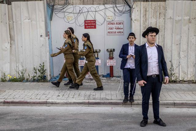 Israeli female conscripts walk past as ultra-Orthodox Jews, who are largely exempt from military service, gather during a protest against conscription at the entrance to Tel Hashomer, the central recruitment centre of the Israeli Army, in Ramat-Gan on April 12, 2026. Israel’s Prime Minister, who depends on his ultra-Orthodox party allies to maintain power, has employed a range of tactics to delay the adoption of legislation that would allow the enlistment of ultra-Orthodox Jews, who are largely exempt from military service. Under a ruling established at the country’s creation in 1948—when the ultra-Orthodox community was small—men who devote themselves full-time to the study of sacred Jewish texts receive a de facto exemption. (Photo by Ilia YEFIMOVICH / AFP) / 