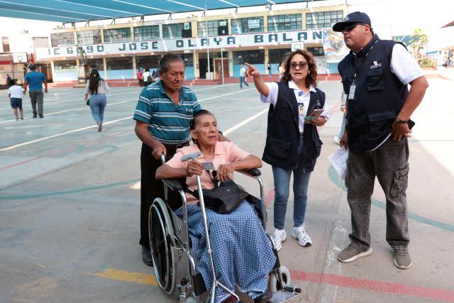 ONPE (National Office of Electoral Processes) employees assist a woman in a wheelchair by helping her find her polling station during the presidential election in Lima on April 12, 2026. Peruvians will elect a new president from a record field of 35 candidates to lead a country plagued by organized crime and chronic political instability. (Photo by Connie FRANCE / AFP)