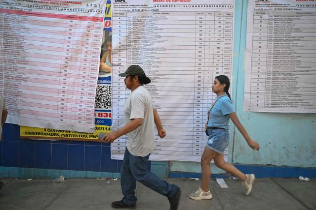 People cross in front of a banner with electoral information at a school polling station in Pamplona Alta, in the San Juan de Miraflores district on the outskirts of Lima, on April 12, 2026. Hours-long delays marred Peru's presidential and legislative elections on April 12, as voters sought to end political chaos that has seen a string of presidents ousted or jailed. (Photo by Ernesto BENAVIDES / AFP)