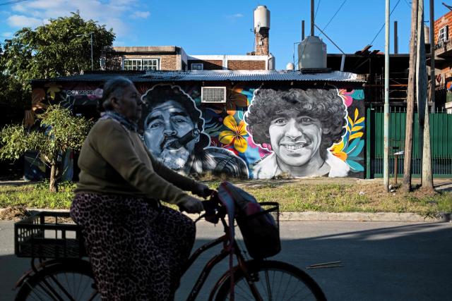 A woman rides a bicycle past murals depicting the late football star Diego Maradona in his hometown neighborhood Villa Fiorito, in Buenos Aires province, Argentina, on April 9, 2026. The second trial over the death of football star Diego Maradona begins on April 14, 2026, after the first one, which took place last year, was annulled when it was discovered that one of the judges was taking part in a clandestine documentary about the proceedings. (Photo by TOMAS CUESTA / AFP)