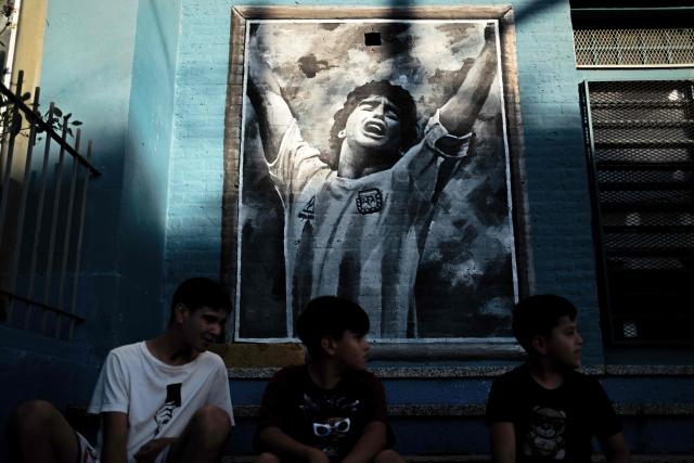 Boys sit in front of a mural depicting the late football star Diego Maradona in his hometown neighborhood Villa Fiorito, in Buenos Aires province, Argentina, on April 9, 2026. The second trial over the death of football star Diego Maradona begins on April 14, 2026, after the first one, which took place last year, was annulled when it was discovered that one of the judges was taking part in a clandestine documentary about the proceedings. (Photo by TOMAS CUESTA / AFP)