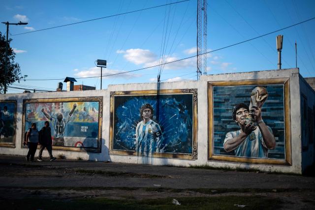 People walk past murals depicting the late football star Diego Maradona in his hometown neighborhood Villa Fiorito, in Buenos Aires province, Argentina, on April 9, 2026. The second trial over the death of football star Diego Maradona begins on April 14, 2026, after the first one, which took place last year, was annulled when it was discovered that one of the judges was taking part in a clandestine documentary about the proceedings. (Photo by TOMAS CUESTA / AFP)