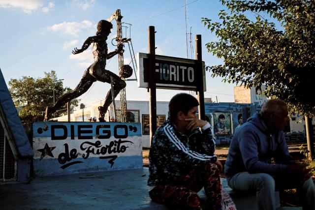 People sit next to a statue depicting the late football star Diego Maradona in his hometown neighborhood Villa Fiorito, in Buenos Aires province, Argentina, on April 9, 2026. The second trial over the death of football star Diego Maradona begins on April 14, 2026, after the first one, which took place last year, was annulled when it was discovered that one of the judges was taking part in a clandestine documentary about the proceedings. (Photo by TOMAS CUESTA / AFP)