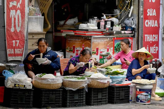 Women sit and eat rice noodles on the sidewalk in Hanoi on April 14, 2026. (Photo by Nhac NGUYEN / AFP)
