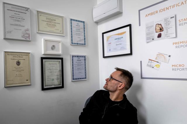 Michal Kosior, gemologist, amber specialist and founder of "Amber Expert", poses for photos in front of several certificates in his laboratory in Gdansk, northern Poland, on March 11, 2026. Formed some 40 million years ago from conifer resins, amber often contains air bubbles, plant fragments or insects trapped before fossilisation, granting each piece a unique "fingerprint". In Gdansk itself, workshops, shops and stalls dedicated to amber abound on every corner of the historic centre, each attracting flocks of tourists. (Photo by Wojtek RADWANSKI / AFP)