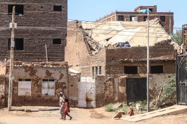 A woman and a child walk past a damaged building in the capital Khartoum on April 15, 2026, on the third anniversary of the start of the war between the army and its paramilitary foes. On the third anniversary of the start of the grinding conflict on April 15, donors will gather in Berlin for an international conference aimed at reviving faltering peace talks and mobilising aid for one of the world's worst humanitarian crises. (Photo by Ebrahim Hamid / AFP)