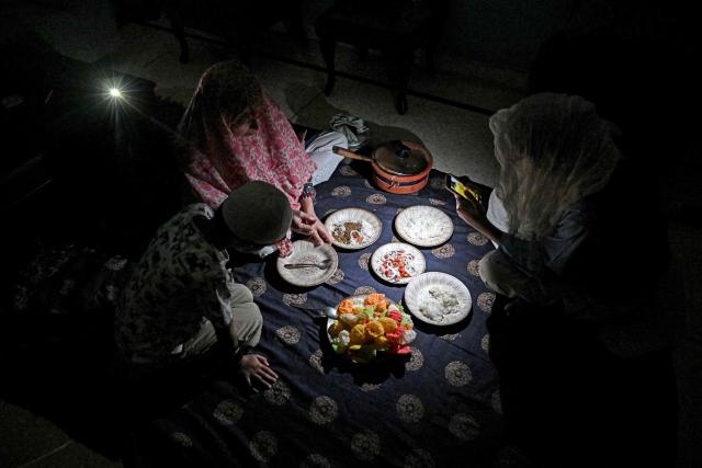 A family eats dinner at home by the light of a mobile phone torch during a power cut in Islamabad on April 15, 2026. Pakistan will suspend electricity supply for about two hours during peak-usage times every evening, the government said, in an effort to manage energy prices affected by the Iran war. (Photo by Aamir QURESHI / AFP)