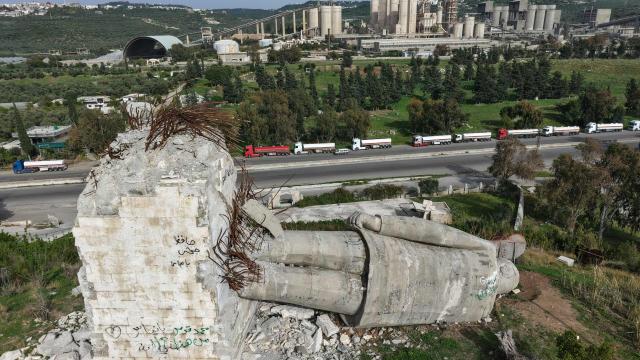 An aerial photograph shows a destroyed statue of the late Syrian president Hafez al-Assad, as a long line of Iraqi diesel-laden tanker trucks line up along the Tartus-Baniyas highway waiting to unload their cargo at the Baniyas port refinery on the Mediterranean Sea, on April 15, 2026. Iraq has begun exporting crude using tanker trucks through Syria, its oil ministry said, as an official said oil revenue last month dropped more than 70 percent compared to February. More than a month into the Middle East war that has wrought havoc on energy markets, Iraq has been particularly impacted as oil exports account for some 90 percent of its budget revenues. Until Iranian attacks and threats all but shut the Strait of Hormuz in revenge for US-Israeli strikes, Iraq exported the majority of its oil through the strategic waterway. (Photo by Bakr ALkasem / AFP)