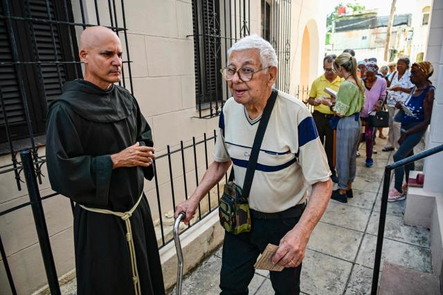 Fray Luis Pernas stands next to an elderly man waiting to receive free medicine as social assistance at the Church of the Holy Cross of Jerusalem in Havana on April 14, 2026. Dozens of people, mainly elderly and low income persons, come to this church to receive free medicines they cannot find in pharmacies or hospitals, reflecting the social role churches are playing in a Cuba in crisis. (Photo by YAMIL LAGE / AFP)